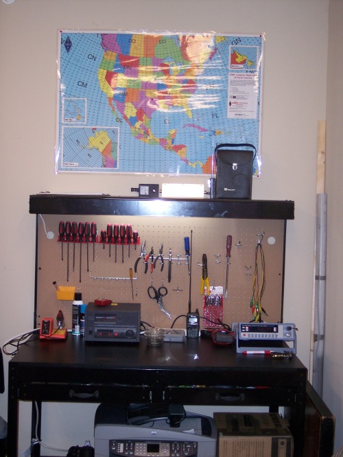 The K5ASP radio shack includes a well equipped work bench for construction projects and repairs. A printer, scanner, fax, and copier unit sits on the bottom shelf of the workbench. A map on the wall above the workbench identifies Canada and United States of America grid squares defined by the Maidenhead Locator System. 